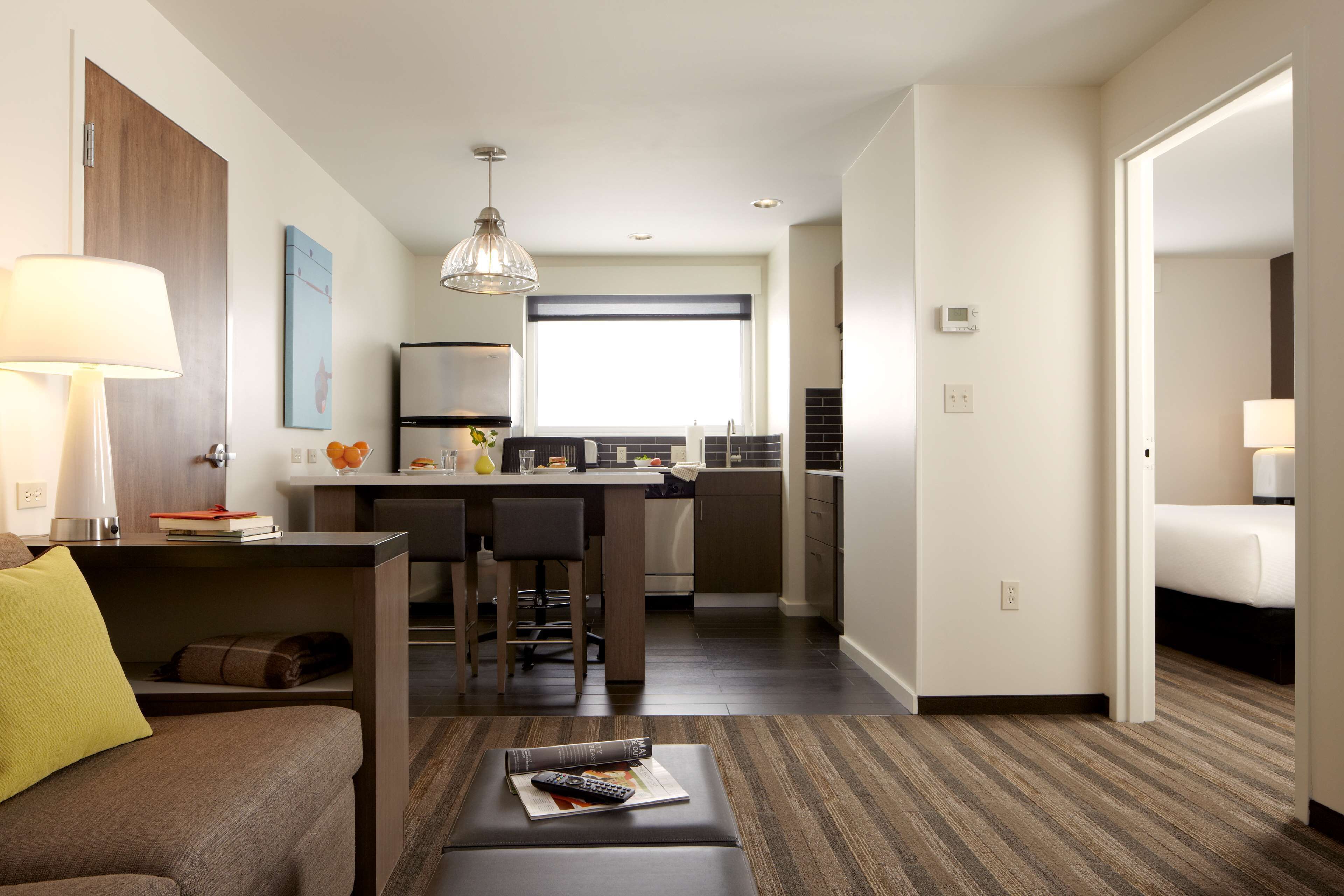A hotel room with a sofa, a coffee table with a remote control, a dining area with a table and chairs, a kitchen area, and a bed.
