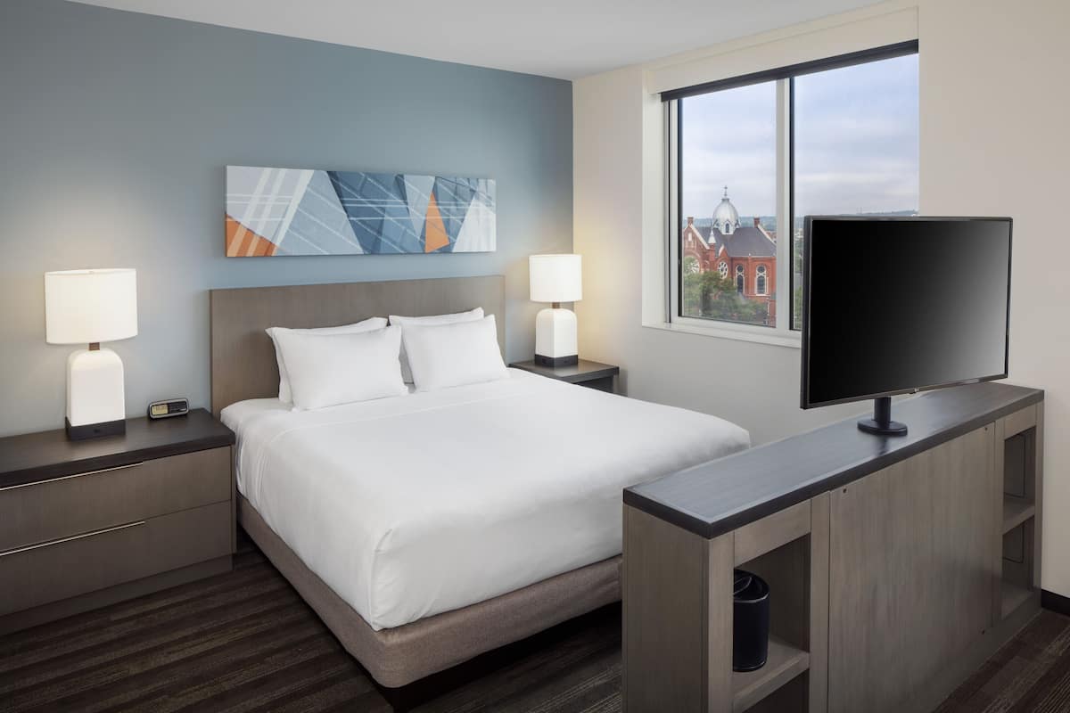 A hotel room with a large bed, two bedside tables with lamps, a flat-screen TV, and a window with a view of buildings.
