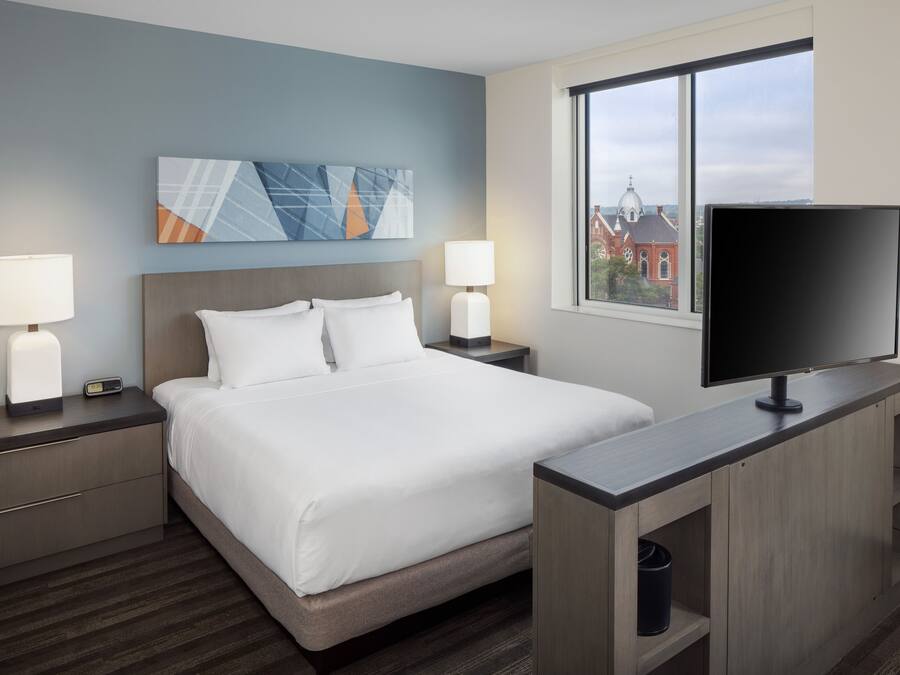 Habitación de hotel con una cama grande, dos mesitas de noche con lámparas, un televisor de pantalla plana y una ventana con vistas a edificios.