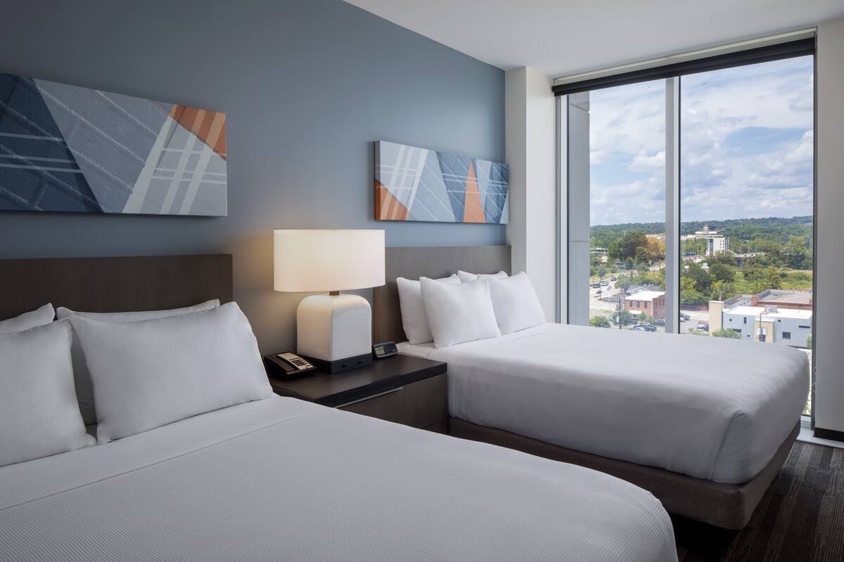 A hotel room with two beds, a nightstand with a lamp, and a large window offering a city view.