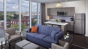 A modern living room with a blue sofa, grey armchairs, and a coffee table. It features a large window with a city view and a kitchen with grey cabinets and stainless steel appliances. - Hyatt House Augusta Downtown (Augusta)