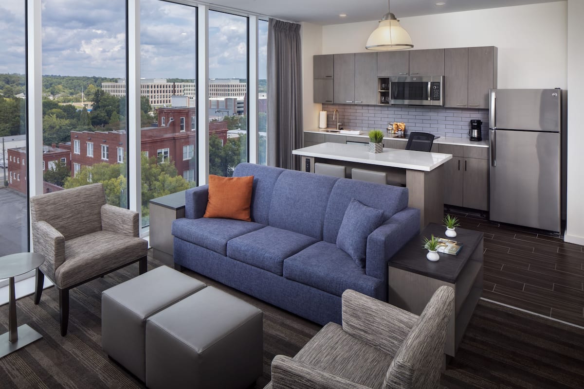 A modern living room with a blue sofa, grey armchairs, and a coffee table. It features a large window with a city view and a kitchen with grey cabinets and stainless steel appliances.