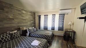 Una habitación con dos camas, un muro de madera, una ventana con cortinas estampadas, un escritorio y una silla.