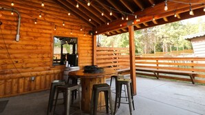 Una cabaña de madera con un porche cubierto que cuenta con una mesa de madera y taburetes tipo bar, luces colgantes y vistas del bosque circundante.