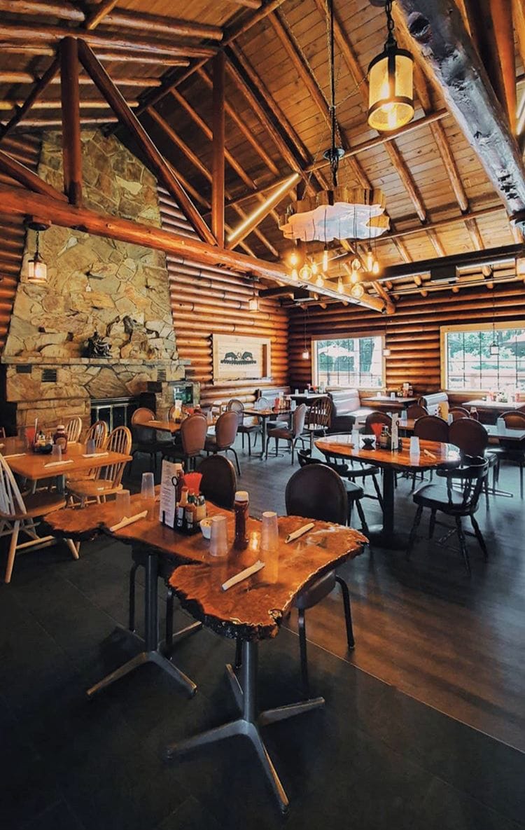 A rustic wooden interior with stone walls, wooden tables and chairs, and a stone fireplace.