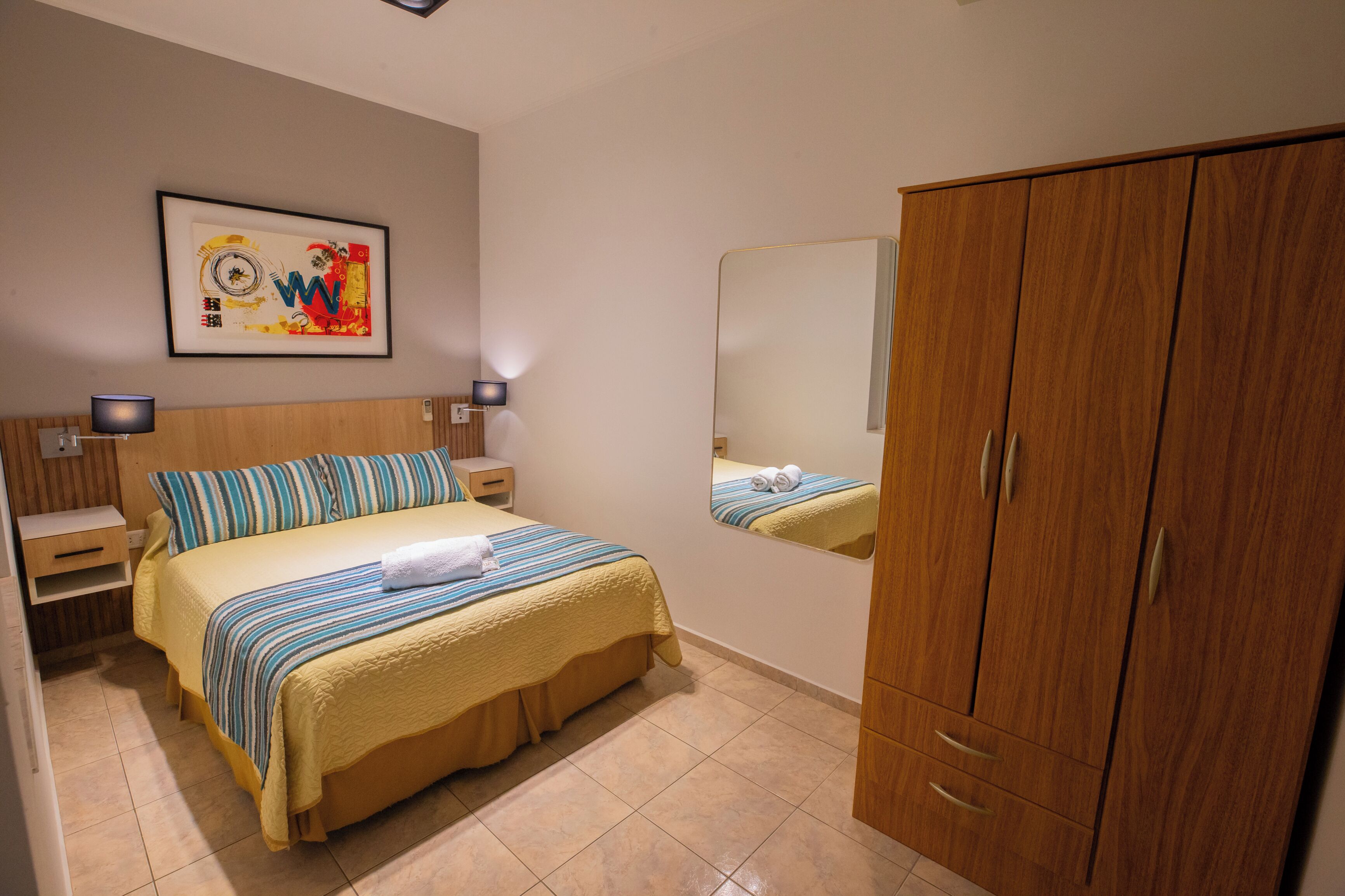 A bedroom with a bed, a nightstand, a wooden wardrobe, a wall-mounted mirror, and a framed artwork.