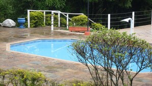 A swimming pool with a tiled deck, surrounded by greenery and a fence.
