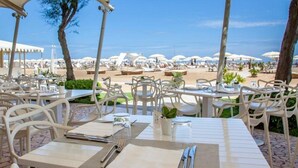 An outdoor dining area with white tables and chairs, overlooking a beach with umbrellas. - Residenza Parco Fellini (Rimini)