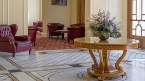 A lobby with a marble floor, red leather chairs, a gold table with a vase of flowers, and a large window with a decorative grille. - Residenza Parco Fellini (Rimini)
