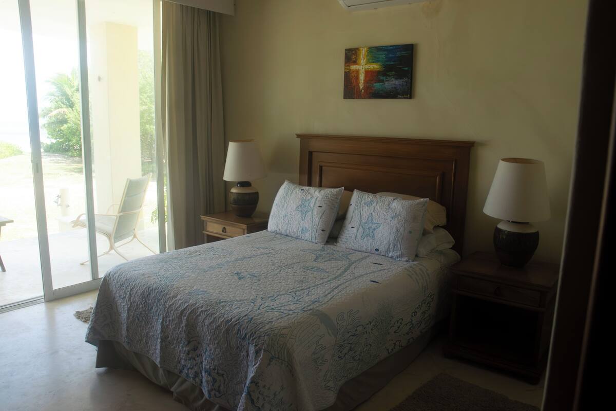 Un dormitorio con una cama grande, dos lámparas de noche, un cuadro con una cruz y una puerta corrediza de vidrio a un balcón.