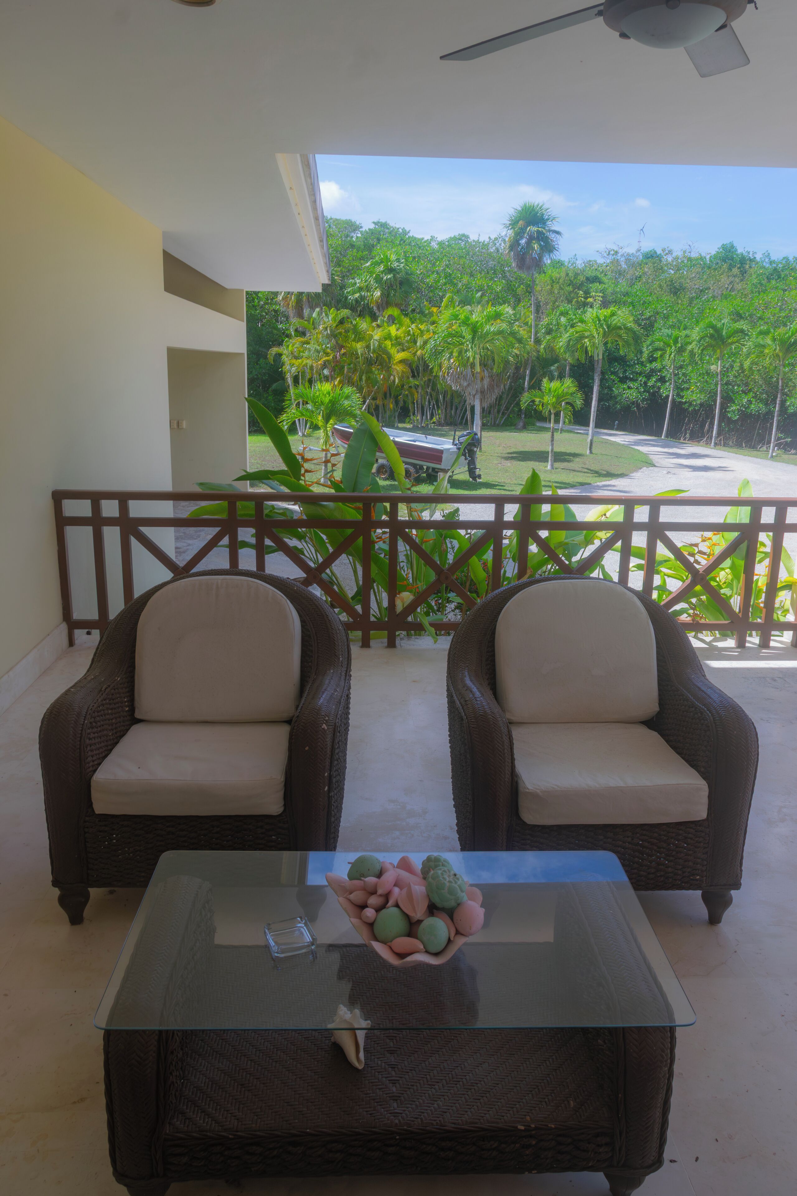 A balcony with wicker chairs and a glass table, overlooking a lush garden with palm trees and a road.