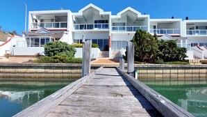 Una propiedad moderna frente al agua con un muelle de madera que conduce a un edificio con varios balcones y grandes ventanales.