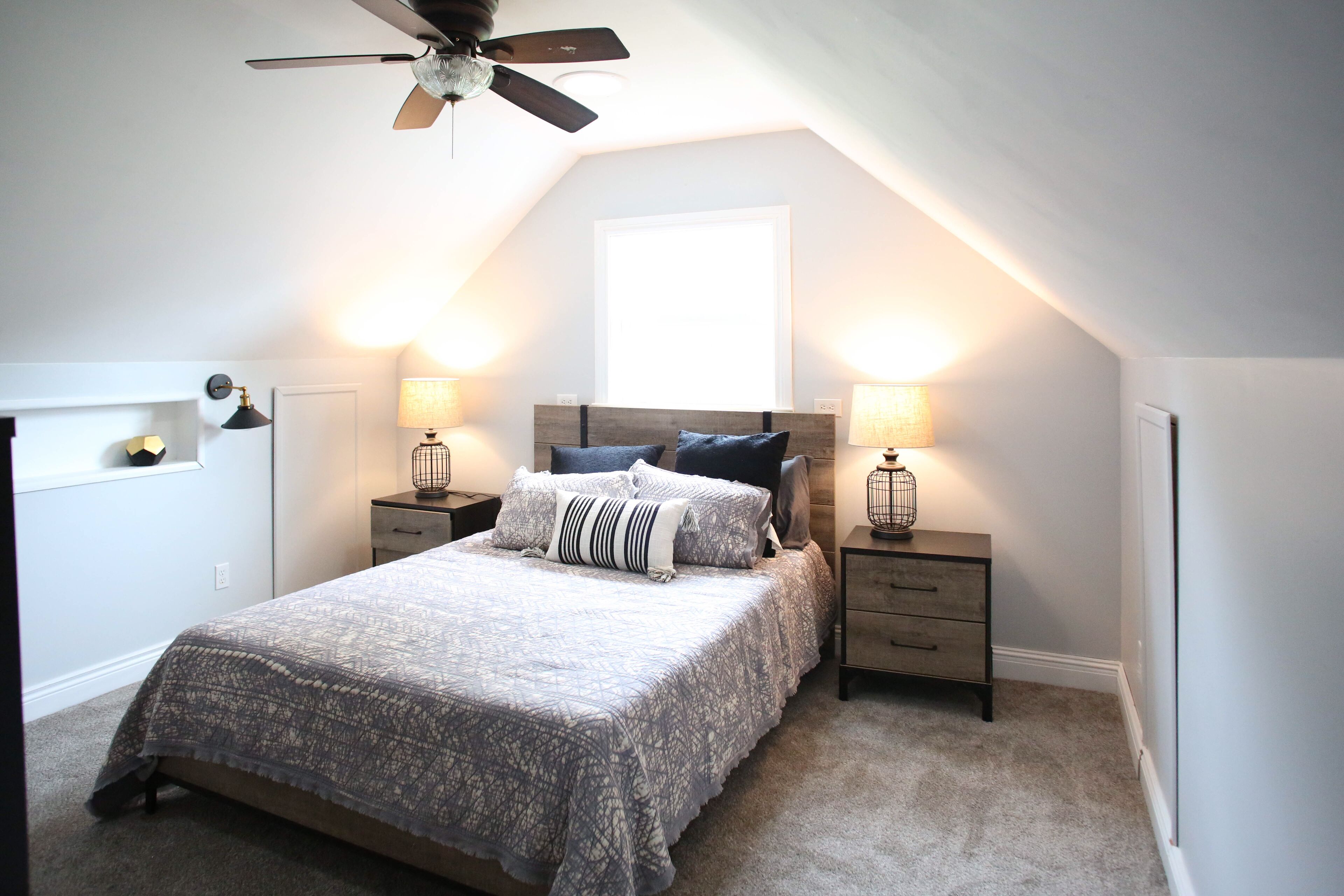 A bedroom with a sloped ceiling, a bed with patterned bedding, two bedside tables with lamps, and a ceiling fan.