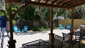Eine überdachte Terrasse im Freien mit Metallstühlen und einem Tisch, umgeben von einer Holzdielenveranda und einer Sandfläche mit blauen Stühlen und einem Holzzaun.