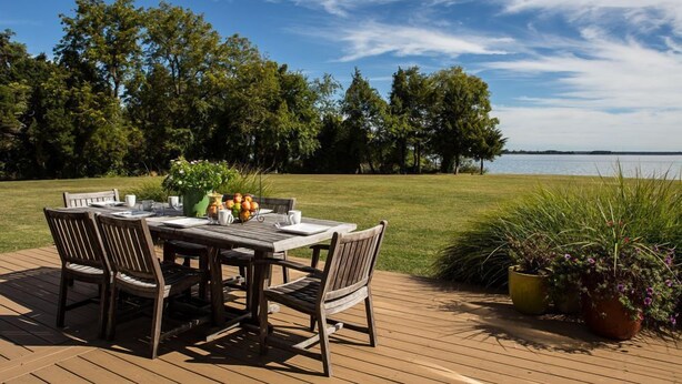 Une terrasse extérieure en bois, avec une table et des chaises, entourée de verdure et offrant une vue sur un plan d’eau.