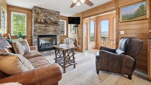 A cozy living room with a stone fireplace, leather sofa, and a ceiling fan. - Amazing View! Sauna Gameroom Firepit Spa Room Rustic Spacious Private Retreat  (Banner Elk)