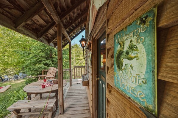 A wooden porch with a welcome sign, outdoor seating, and a view of a lush garden.