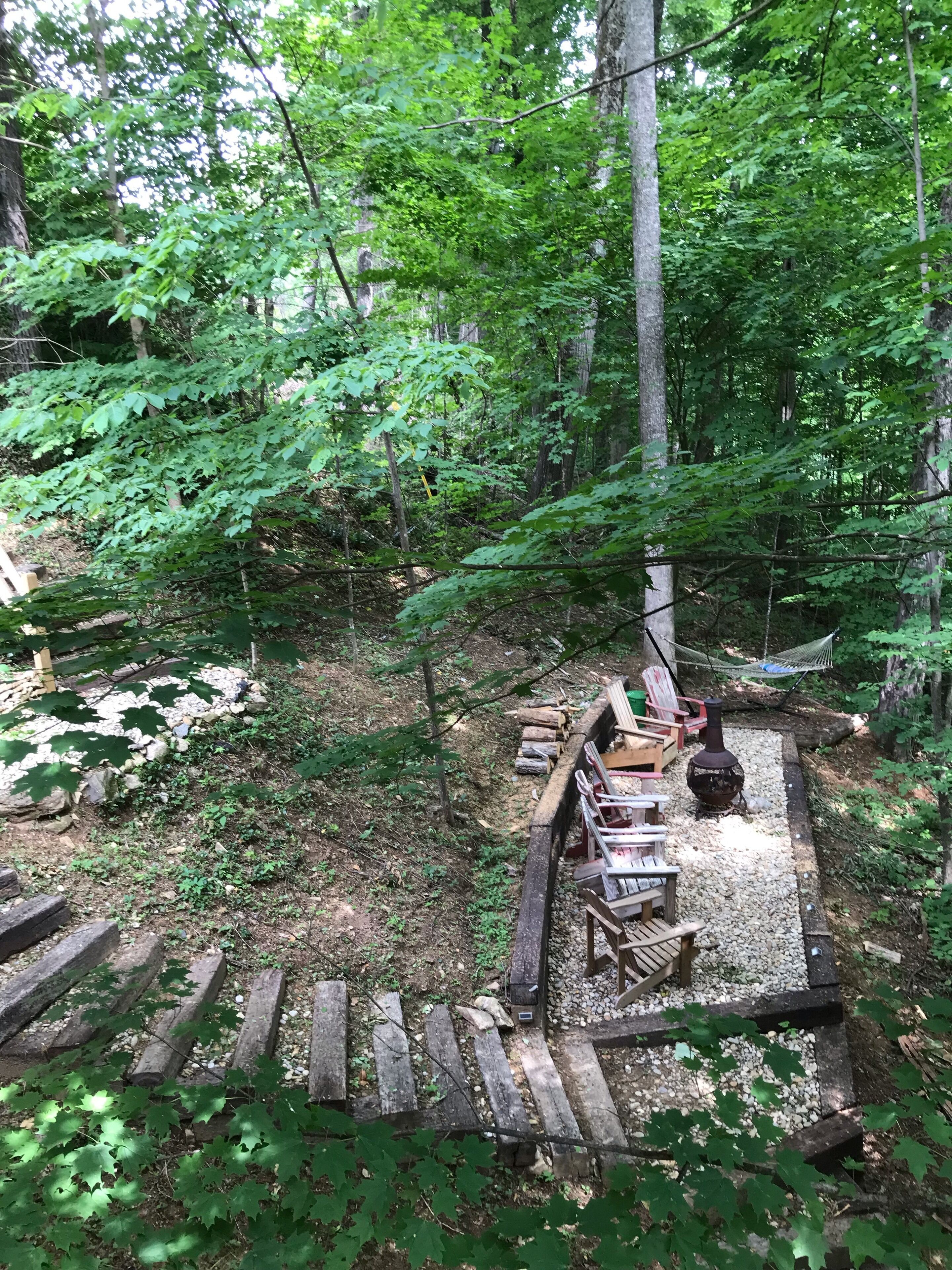 Un espace extérieur aménagé dans un style rustique, avec des bancs en bois et un foyer en pierre, le tout entouré d’une clôture en bois.
