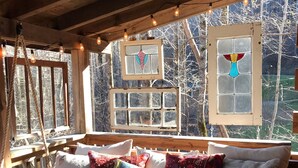 A cozy cabin interior with a wooden bench and a cushioned seating area, large windows with decorative glass panes, and string lights. - ♥♥ Log cabin tree top setting with fire pit!  Sleeping porch. Easy Access (Maggie Valley)