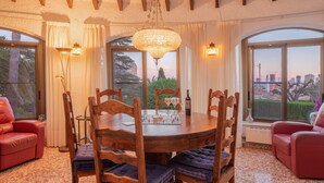 A dining area with a wooden table, chairs, and a chandelier. There are two red sofas and a large window with a view of the city. - Well maintained charming villa**** near the sea Lots of privacy, separate studio (Calp)