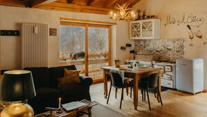 A cozy living room with a wooden ceiling, a dining area with a table and chairs, a kitchen with a stove and refrigerator, and a large window with a view of trees.