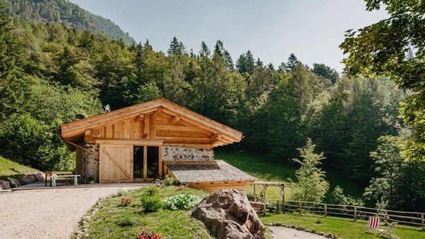 A wooden cabin with a stone wall, surrounded by a gravel path, a wooden fence, and a large rock.
