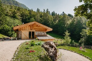 A wooden cabin with a stone wall, surrounded by a gravel path, a wooden fence, and a large rock.