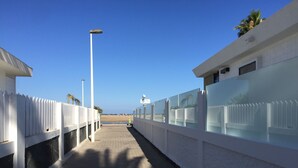 A pathway flanked by white walls with glass panels, leading to a beach.