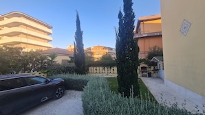 Un patio con un coche, altos cipreses, un seto y un edificio con balcón.