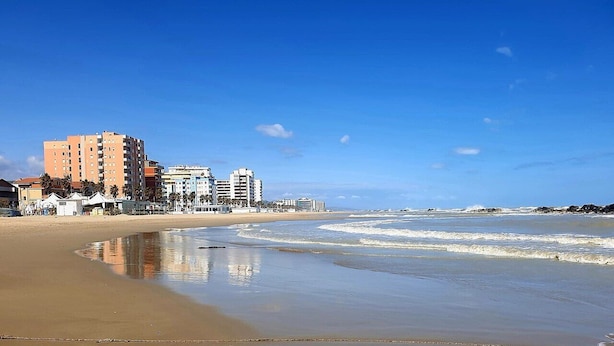 Una playa con arena, un cielo azul despejado y edificios al fondo.