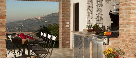 An outdoor dining area with a view of a hilly landscape, a brick wall, and a grill station.