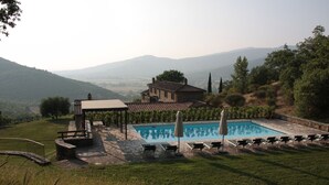 Zona de piscina con sillones de descanso y un pabellón, rodeada por una terraza de piedra y vistas a colinas.