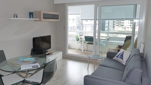 A modern living room with a flat-screen TV on a glass table, a grey sofa, and a balcony with outdoor furniture. - La Marina, Normandy, Apartment 4 people with sea view and port (Courseulles-sur-Mer)