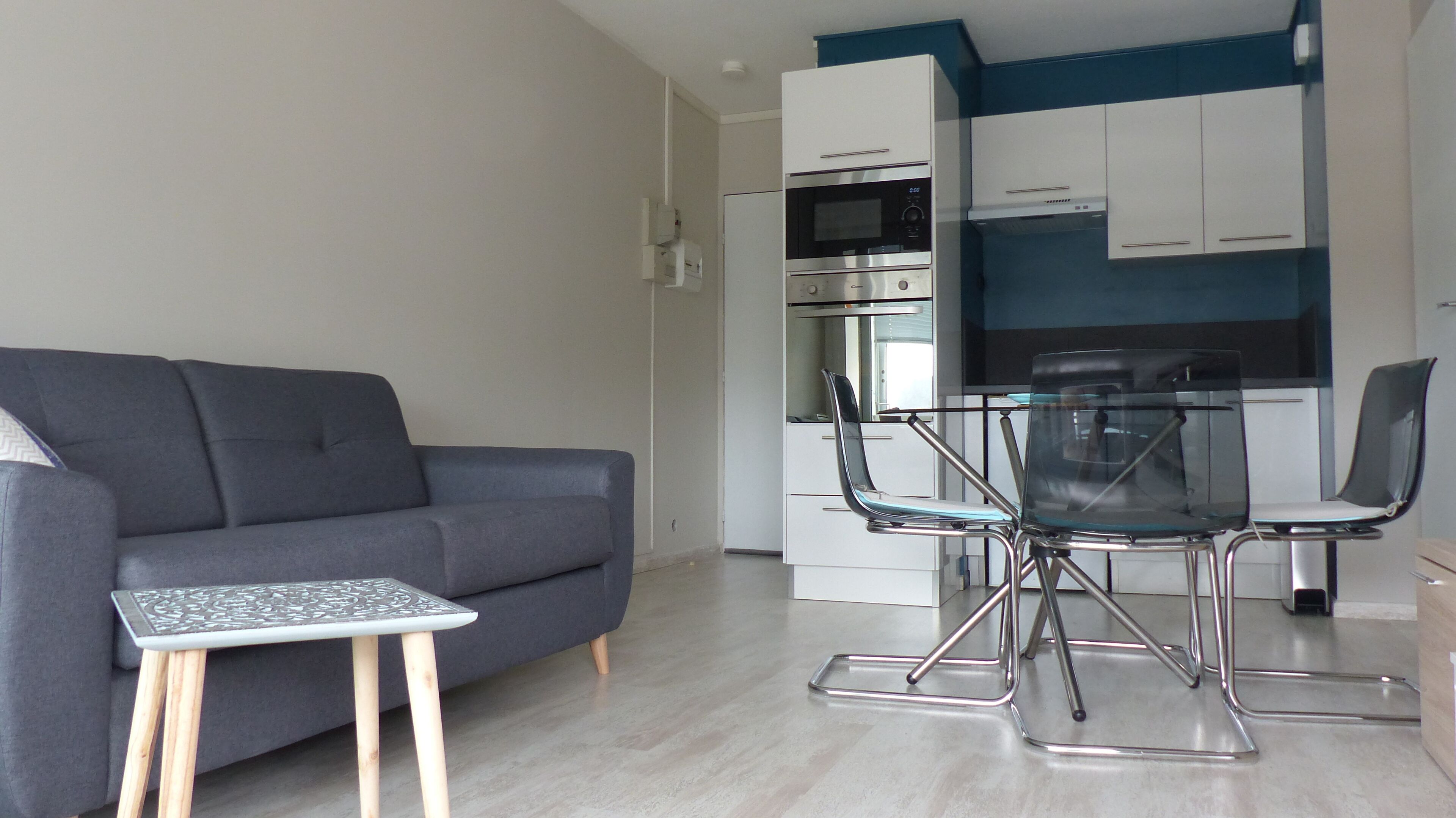 A modern living room with a grey sofa, a small table, and a glass dining table with chairs. A kitchen with white cabinets and a blue accent wall is visible in the background.