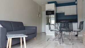 A modern living room with a grey sofa, a small table, and a glass dining table with chairs. A kitchen with white cabinets and a blue accent wall is visible in the background. - La Marina, Normandy, Apartment 4 people with sea view and port (Courseulles-sur-Mer)