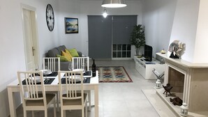 Ein Essbereich mit einem Tisch für vier Personen, einem grauen Sofa mit Kissen, einem großen Fenster und einem weißen Kamin mit Dekorationen.