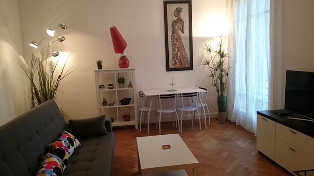 Una sala de estar con un sofá gris, una mesa de centro blanca, una mesa de comedor con sillas, un televisor sobre un mueble bajo y una estantería con objetos decorativos.