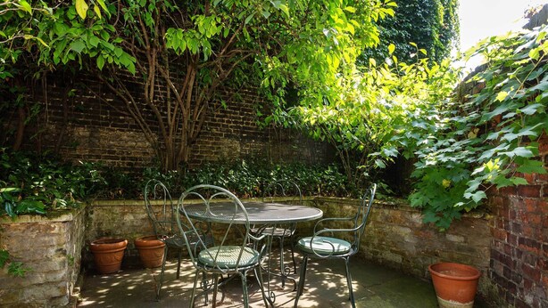 Un mur de briques recouvert de plantes grimpantes, une table ronde en métal avec des chaises et des pots en terre cuite.