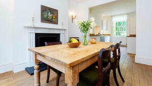 Une salle à manger avec une table en bois, des chaises, un vase de fleurs, un panier de fruits et un tableau au mur.