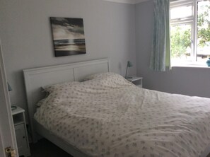 Une chambre à coucher avec un lit, une table de chevet, une lampe, une fenêtre avec des rideaux et un tableau au mur.
