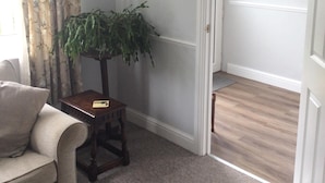 Een kamer met een beige bank, een klein houten tafeltje met een plant erop, en een deur die naar een andere kamer leidt.