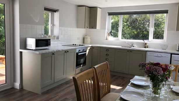 Een moderne keuken met een ingebouwde oven, magnetron en vaatwasser, grote ramen en een eethoek met een tafel gedekt voor twee personen.