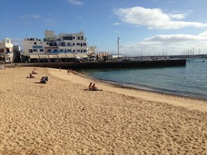 A sandy beach with people sunbathing and walking. There are buildings in the background and a pier with boats. - Casa Patio - Large vacation apartment with terrace and patio by the sea (Corralejo)