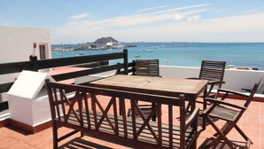 Eine Terrasse mit Tisch und Stühlen, die auf eine Küstenlandschaft mit Booten und einem entfernten Berg blickt.
