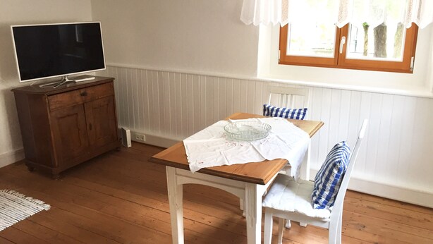 A room with a wooden floor, a white table with a glass bowl, a white chair with a blue and white checkered cushion, a wooden cabinet with a TV on top, and a window with white curtains. - Romantic, listed holiday apartment in Freiburg-Herdern on the church square (Nord)