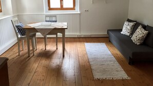 A room with a wooden floor, a dining table with chairs, a sofa with patterned cushions, a window with sheer curtains, and a small shelf on the wall. - Romantic, listed holiday apartment in Freiburg-Herdern on the church square (Nord)