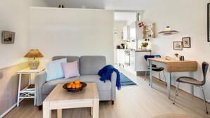 A modern living room with a grey sofa, a small wooden table with fruit, and a dining area with a wooden table and chairs.