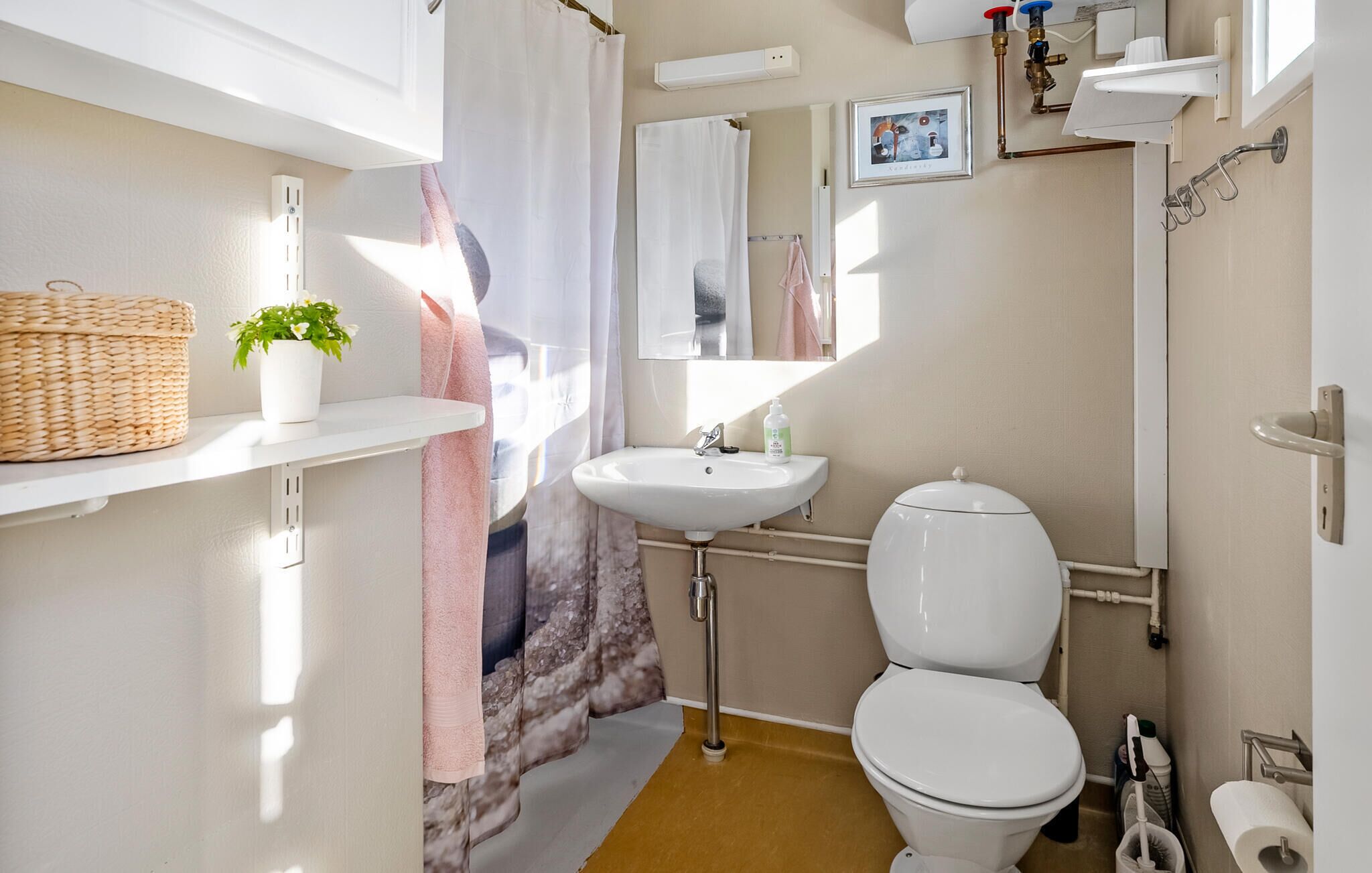 Een badkamer met een witte wastafel, toilet en douchegordijn. Er is een plank met een mand en een potplant.