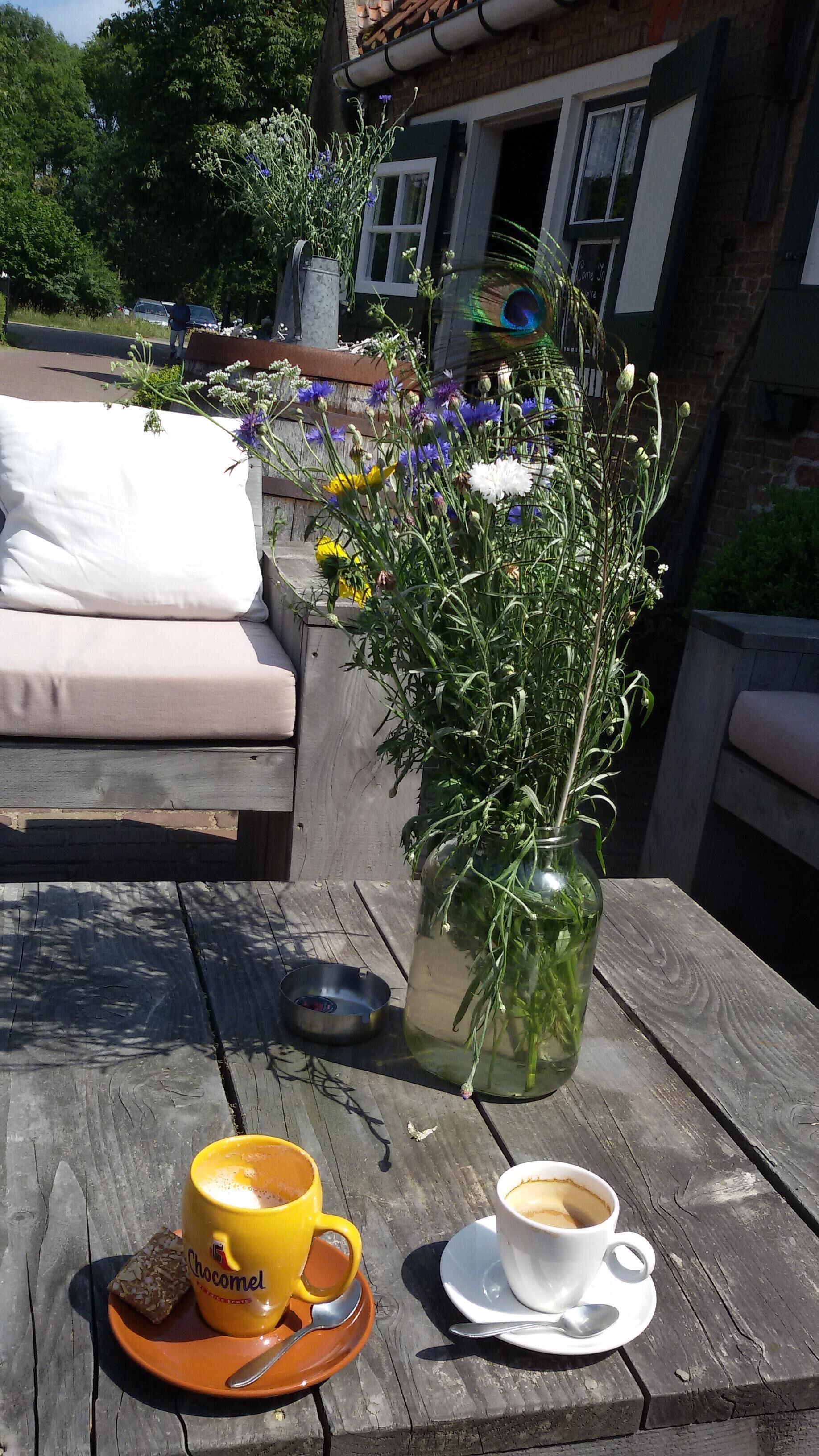 Una mesa de madera con una taza de café, un vaso de Chocomel y un aperitivo en una bandeja. En el fondo, un jarrón con flores.