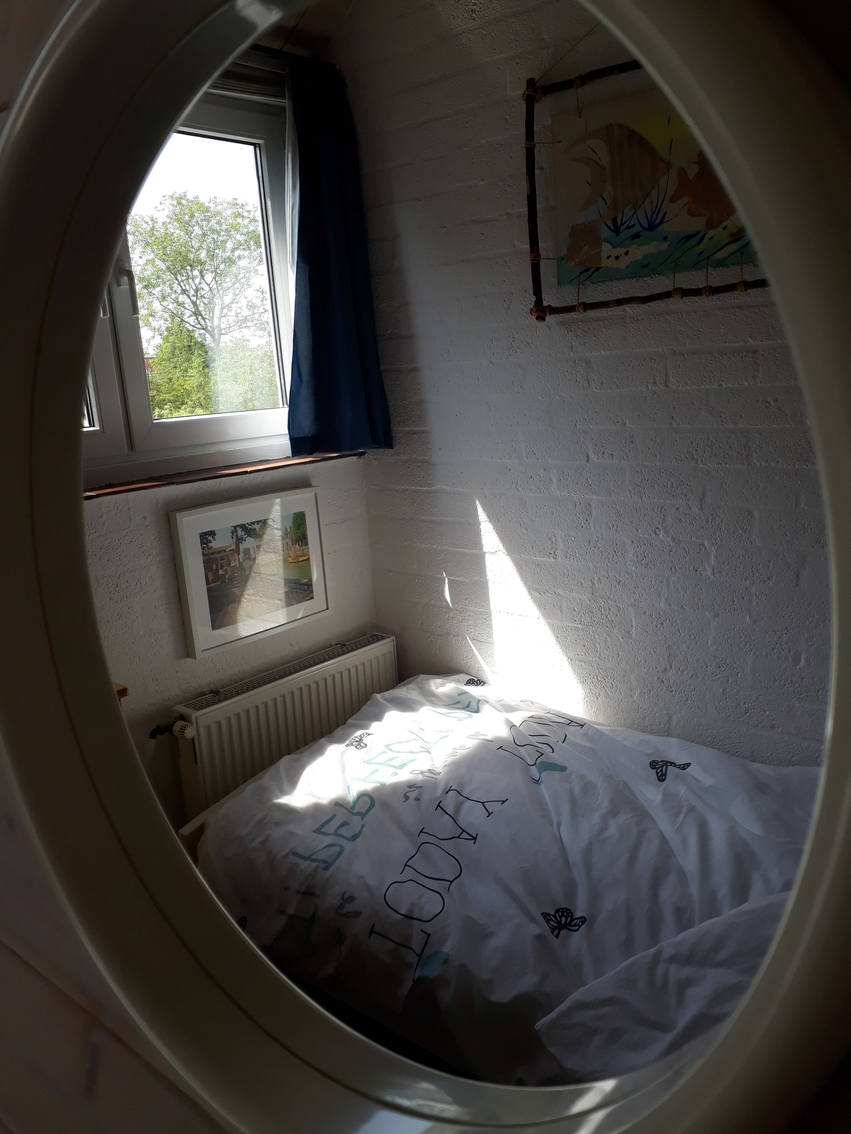 A bedroom with a window, a radiator, a bed with a white sheet, and a framed picture on the wall.